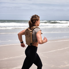 A woman runs on a sandy beach with the Ruck Plate Carrier 3.0 backpack, featuring ergonomic lumbar support, as waves crash in the background.