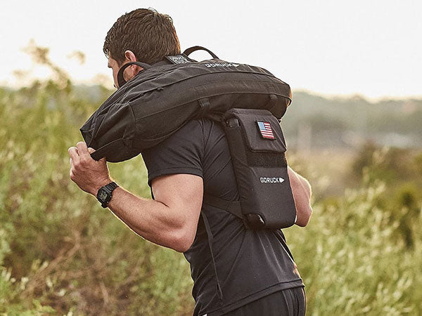 A person outdoors carries a sandbag over their shoulders and wears a black backpack with a U.S. flag patch.