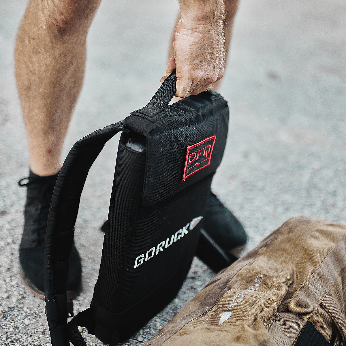 Person holding a black GORUCK backpack next to a brown bag, with visible legs and gravel ground.
