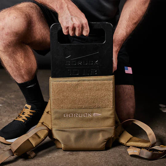 A person inserts a 10 lb GORUCK weight plate into a tan GORUCK backpack on the floor.
