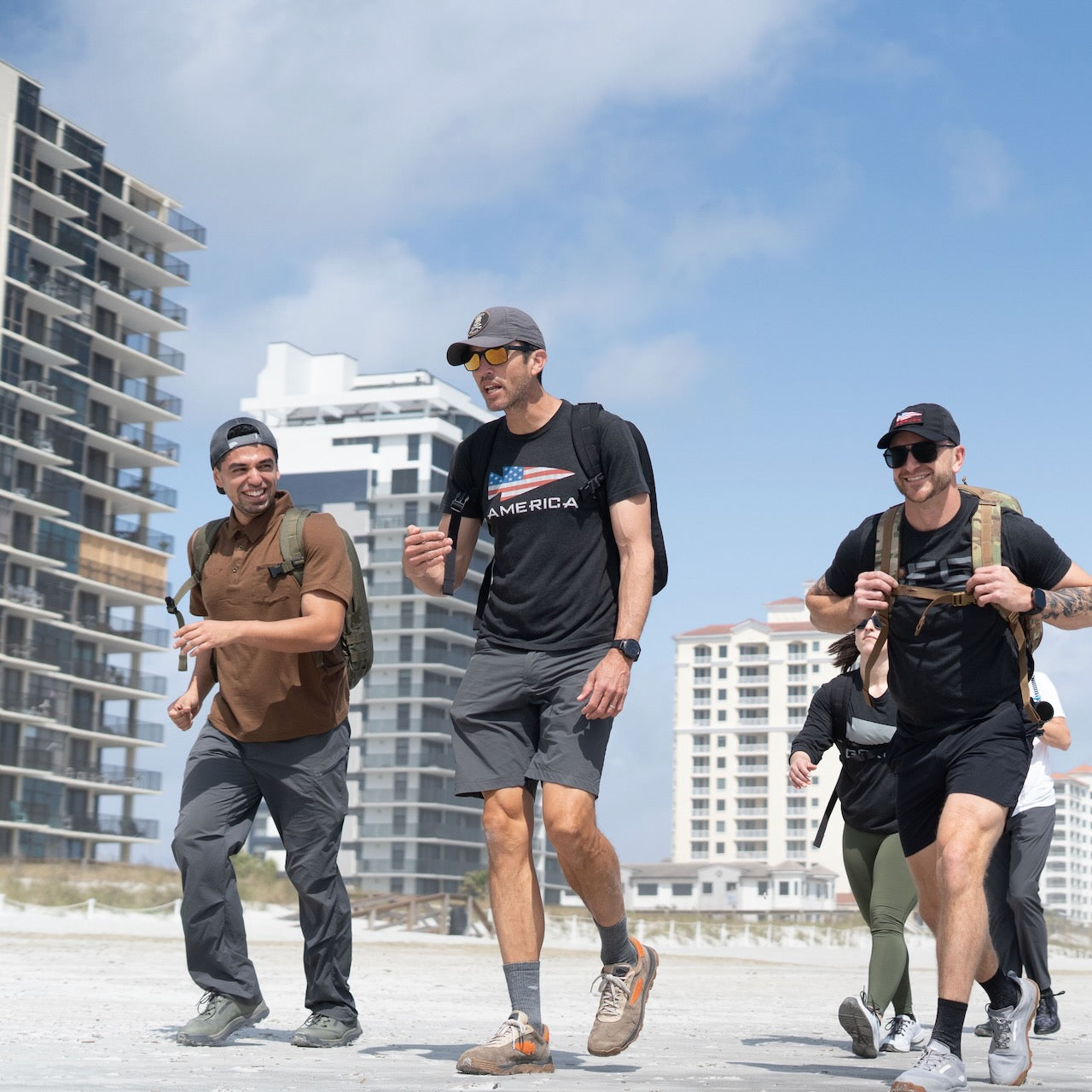 Three people carrying Mackall 45 Fog backpacks walk on a beach near tall buildings, smiling and enjoying the sunny weather.