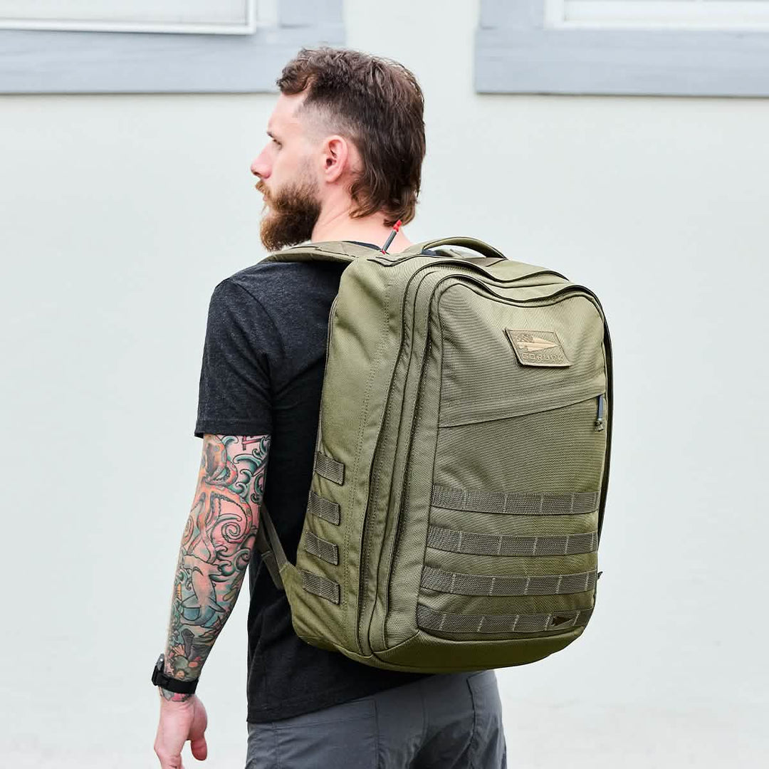 Man with a beard and tattooed arm wearing the GORUCK GR2 large, carry-on compliant travel rucksack, standing sideways against a pale wall.