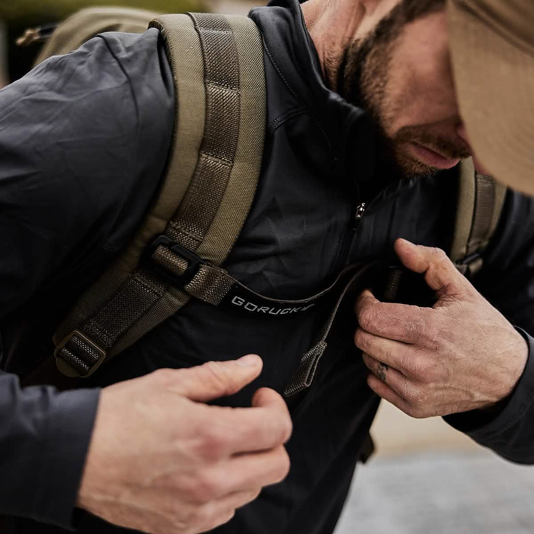 A man with a backpack fastens the chest strap, preparing for rucking with Ruck Plates in his GORUCK Rucker 4.0; only his upper body is visible.