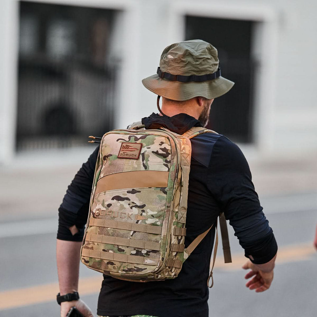 A person wearing a GORUCK Boonie Hat - Tactical - ToughDry and a camo backpack strolls outdoors on a street near a white building.