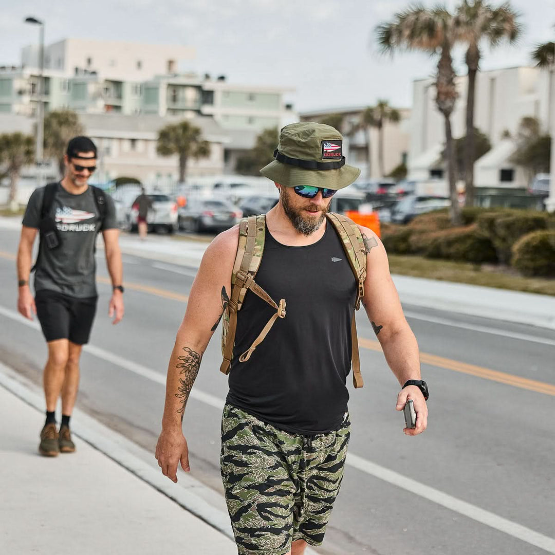 A man in camo shorts and a GORUCK Boonie Hat - Tactical - ToughDry (50+ UPF) walks on the sidewalk, followed by another man; palm trees and buildings in the background.