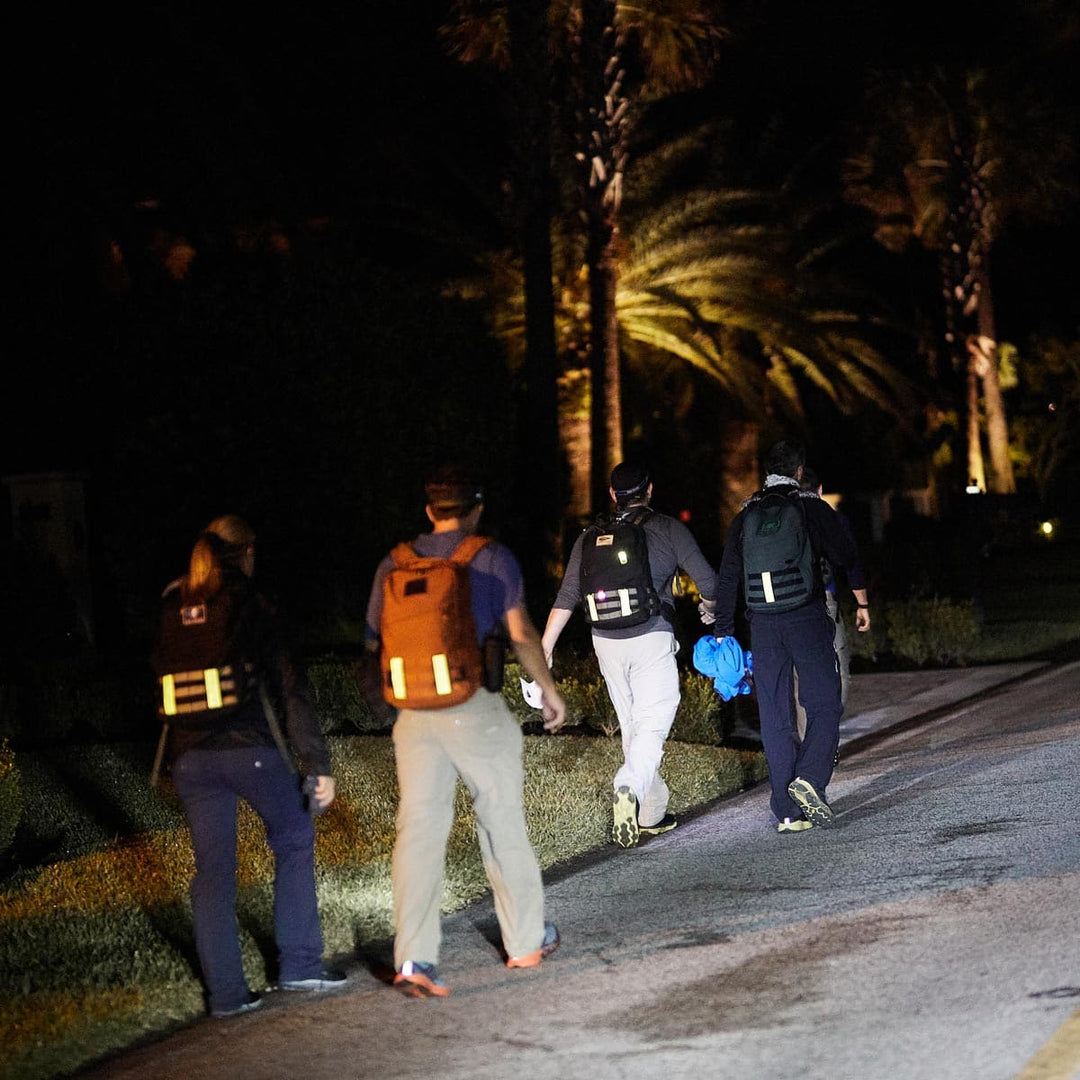 Four people with backpacks, featuring GORUCK's Reflective Ruck Bands for enhanced nighttime visibility, stroll along a dimly lit street.