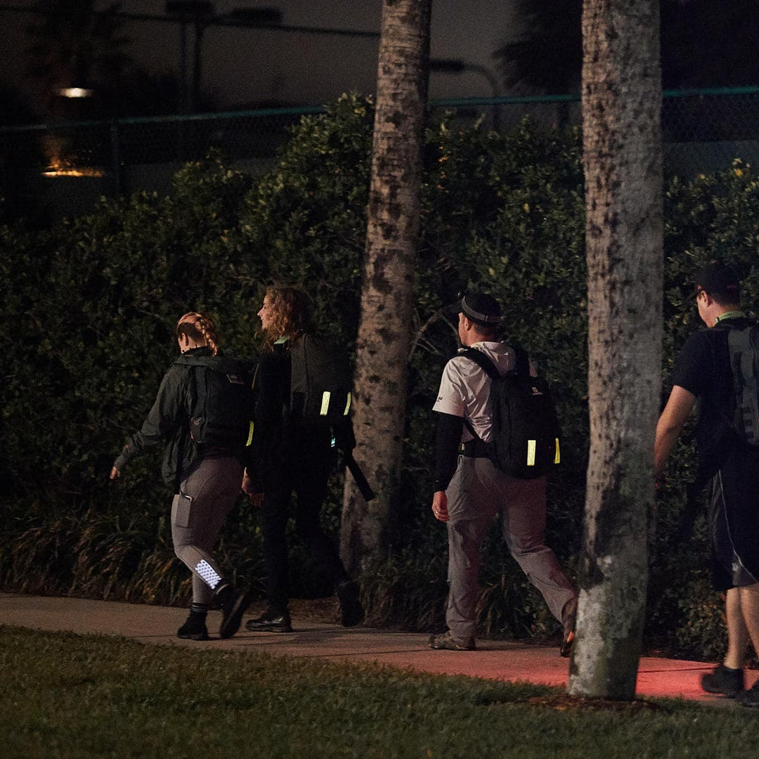 A group of people strolls along a moonlit path at night, their backpacks equipped with GORUCK Reflective Ruck Bands for improved nighttime visibility, surrounded by the silhouettes of trees and bushes.