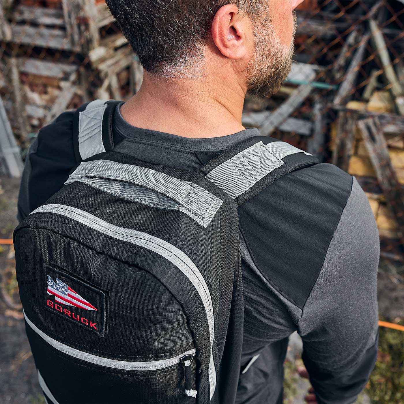 Man wearing a black and gray backpack with an American flag patch, standing by a fence with wooden debris.
