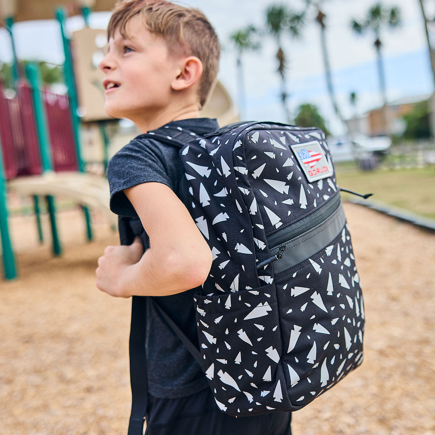 Boy with a black and white patterned backpack standing on a playground with palm trees in the background.