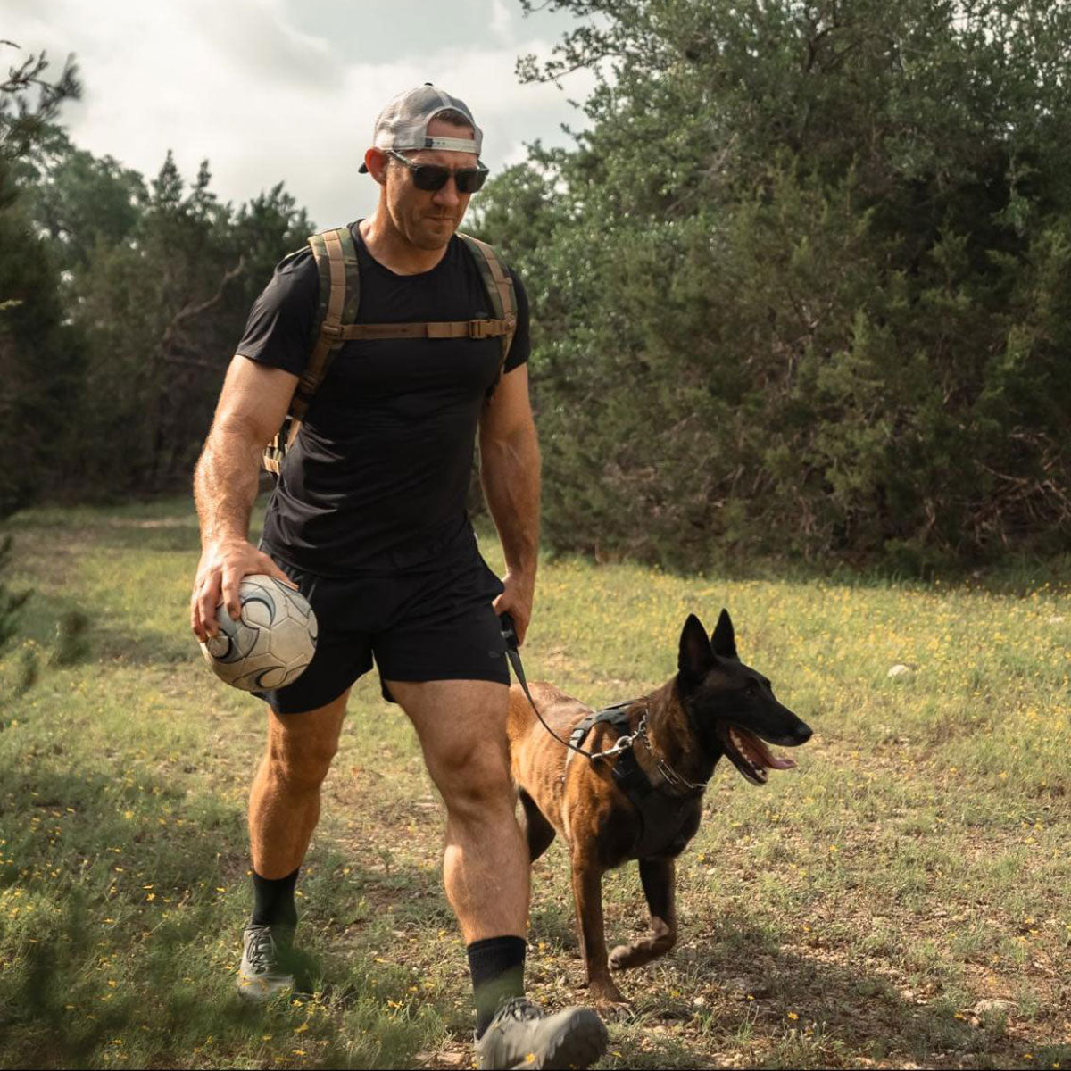 Man in sports attire walks a Belgian Malinois dog on a leash outdoors, holding a soccer ball and wearing sunglasses.