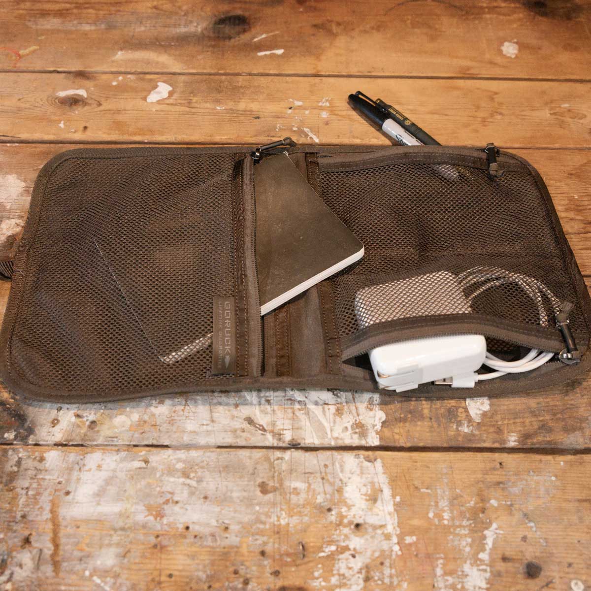 An open mesh travel pouch holds a notebook, cables, and a charger on a worn wooden surface.