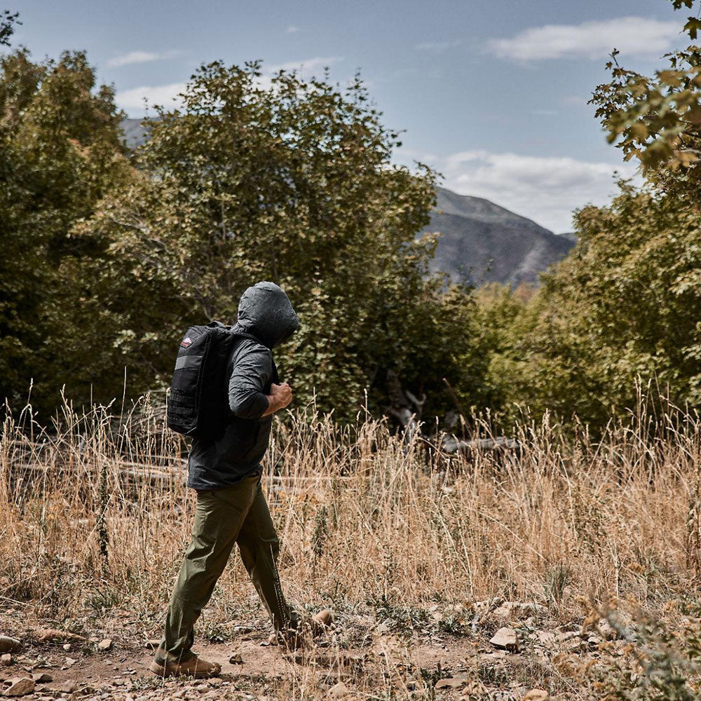 A person in a hooded jacket hikes through a dry, grassy area with trees and mountains in the background.