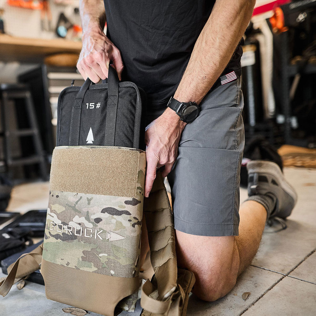 Person kneeling, placing a 15 lb weighted plate into a camouflage vest in a garage setting.