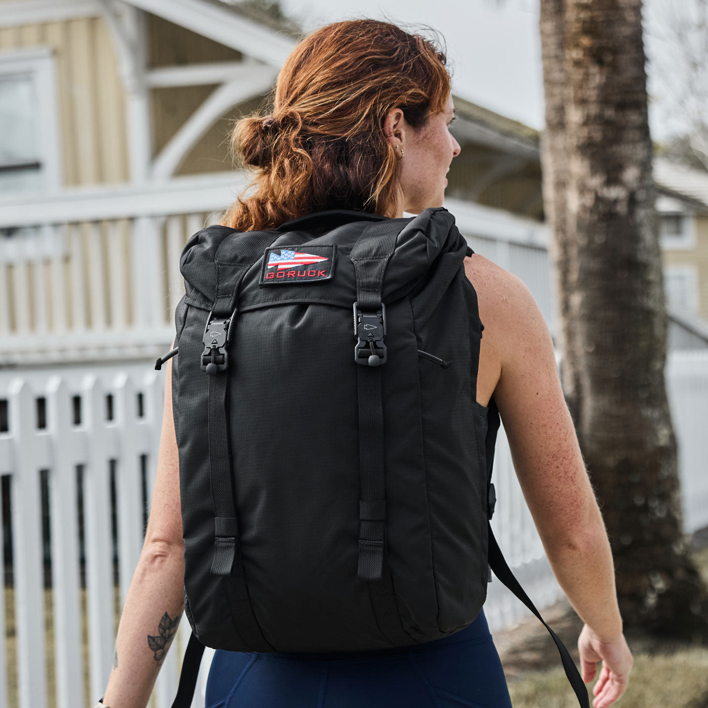 A woman with red hair carries a large black backpack outdoors near a white fence and a tree.