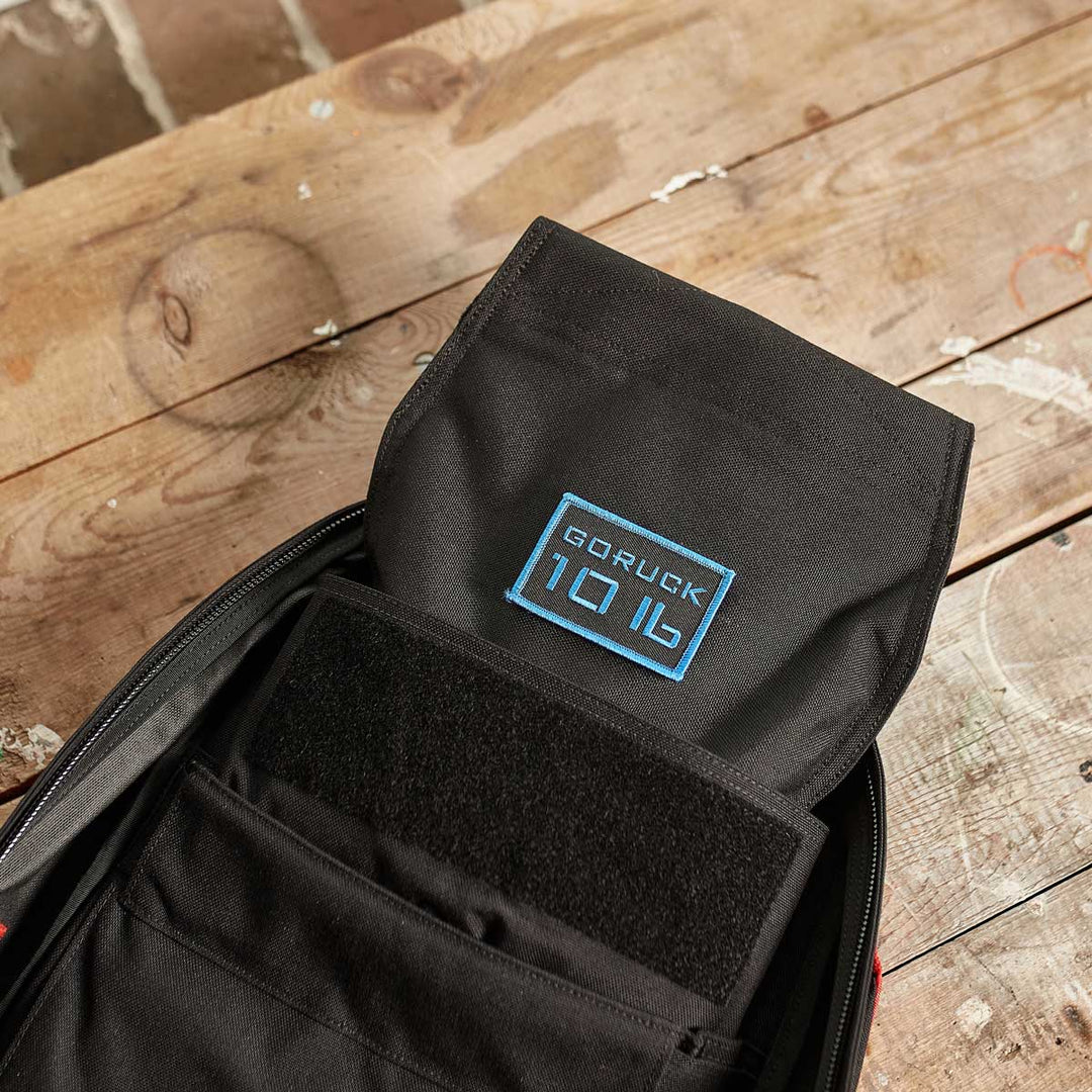 Open black backpack with a visible GORUCK 10L16 patch rests on a rustic wooden surface—ideal for carrying your GORUCK Ruck Sandbag - 10LB (4.5KG) or other workout essentials.