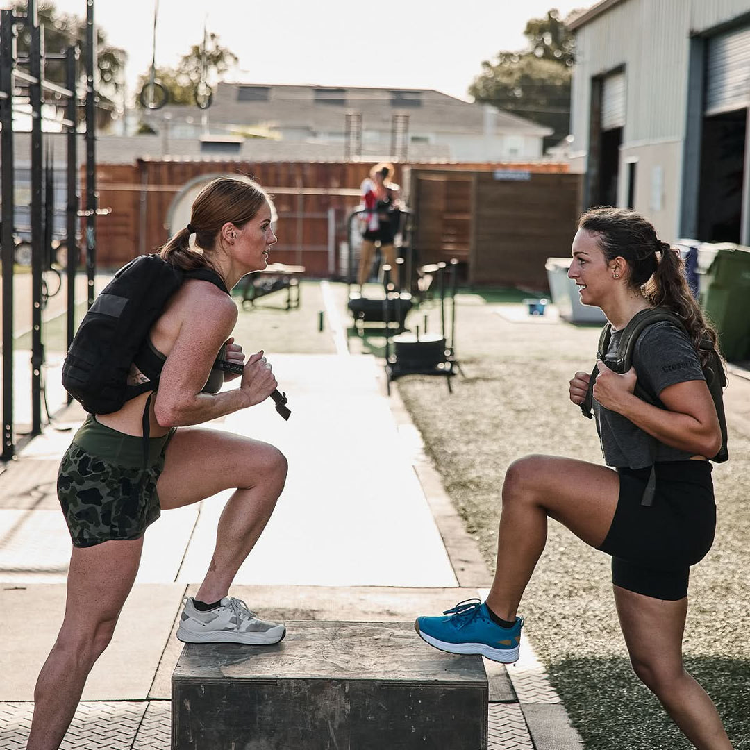 Two women in workout gear, each carrying the Ruck Clubs Starter Kit - Light by GORUCK, doing step-ups outdoors and smiling at each other.