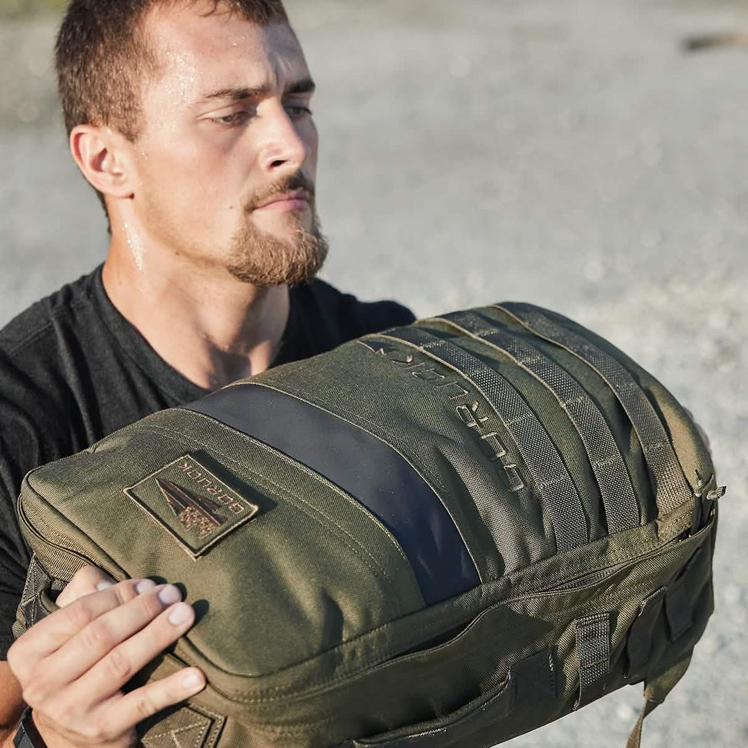A determined man hoists a GORUCK Rucker 4.0 outdoors, focused and sweaty as he embraces the challenge of rucking.