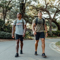 Clad in t-shirts and shorts, two men, who might be utilizing the GORUCK Ruck Clubs Starter Kit -Light, traverse a tree-lined road in an outdoor environment as they fully engage in the sport of rucking.