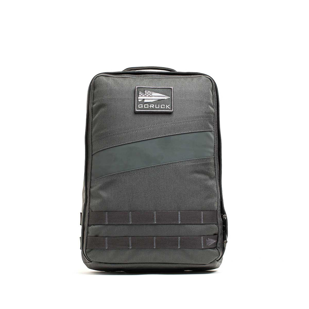 A gray GORUCK Rucker 4.0 fitness rucksack with a patch and black horizontal front straps, standing upright.