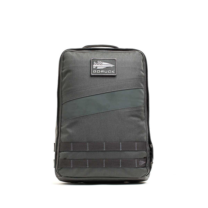 A gray GORUCK Rucker 4.0 fitness rucksack with a patch and black horizontal front straps, standing upright.