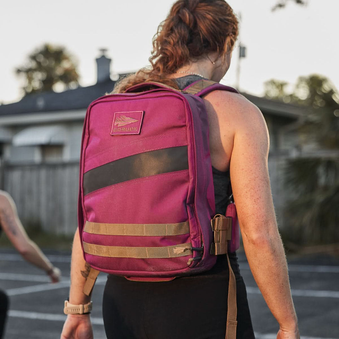 A woman with a ponytail, seen from behind near a house outdoors, wears a magenta GORUCK Rucker 4.0 rucksack.