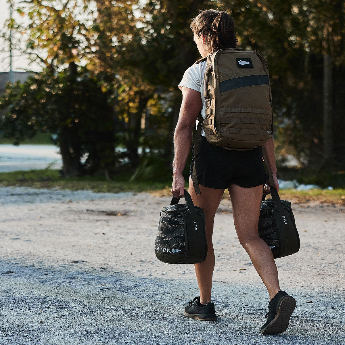 A woman carrying a large backpack and two kettlebells walks outdoors on a gravel path.