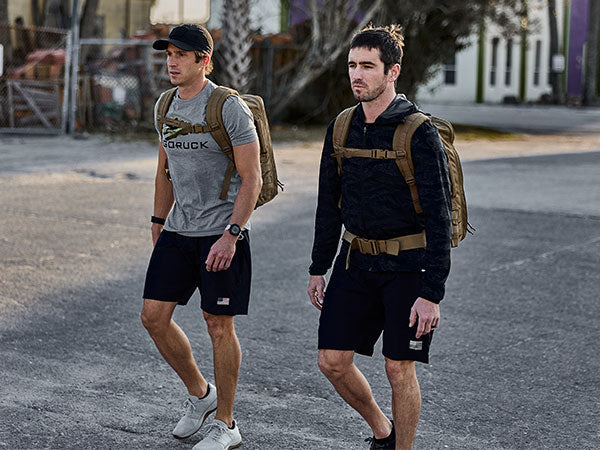 Two men wearing backpacks and athletic clothes walk outdoors on a paved surface with buildings in the background.