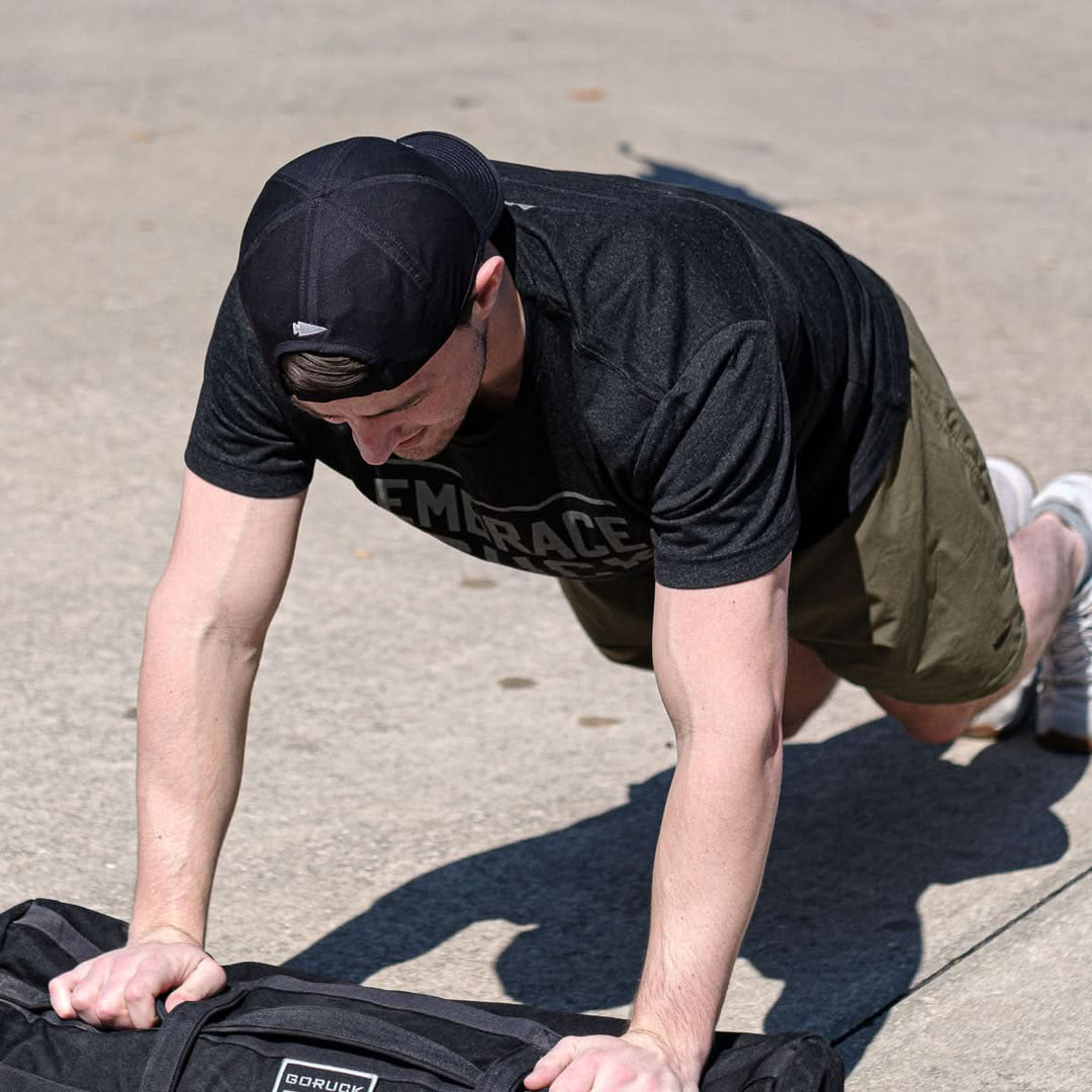 A man wearing the GORUCK Performance Running Hat - ToughMesh and a premium t-shirt does incline push-ups outdoors on a sunny day.