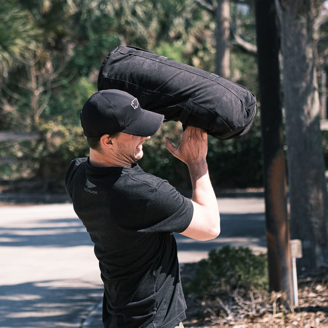 Outdoors, a man lifts a black sandbag over his shoulder while wearing the GORUCK Performance Running Hat - ToughMesh and a black shirt.
