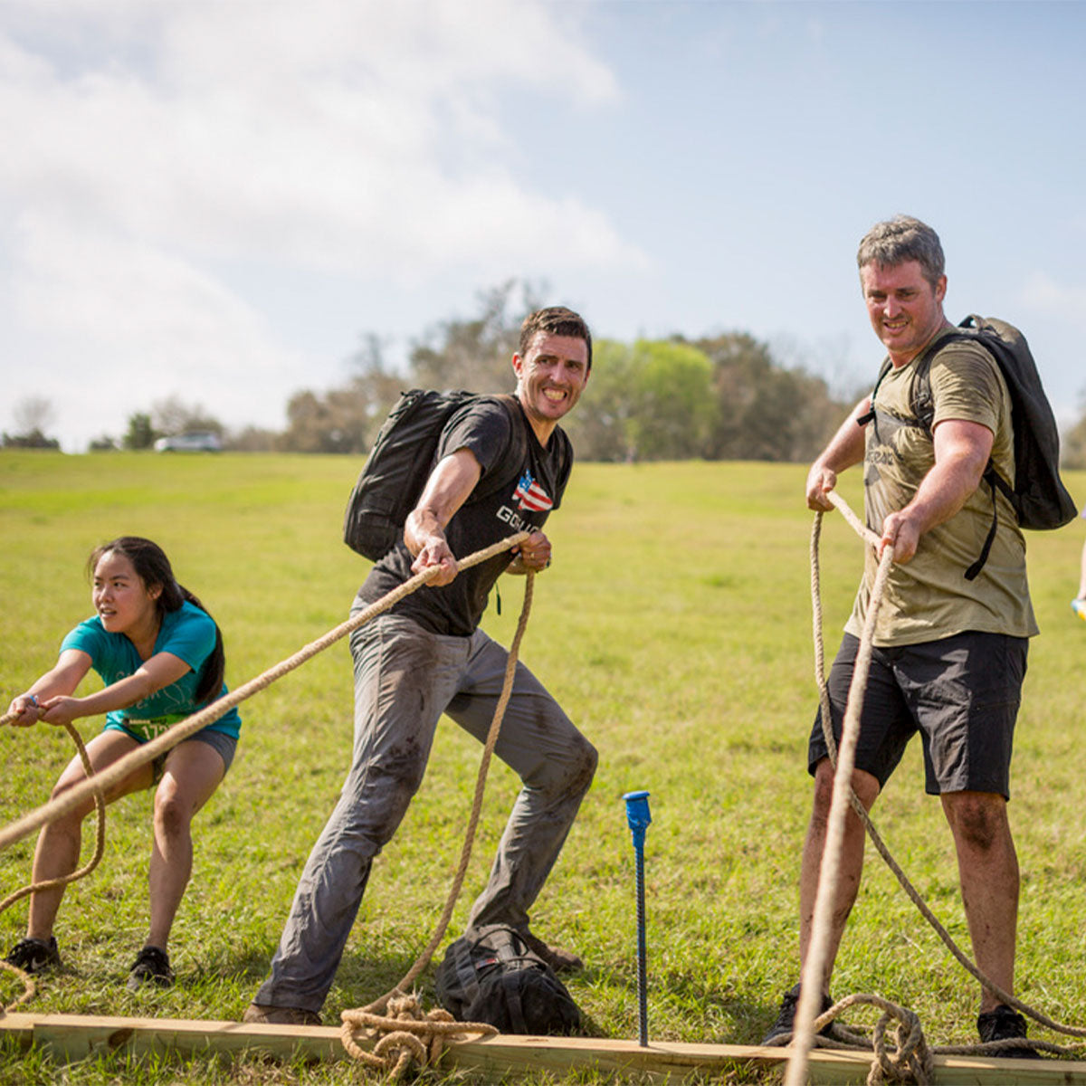 Three people outdoors, smiling and pulling ropes together in a teamwork activity on a grassy field.