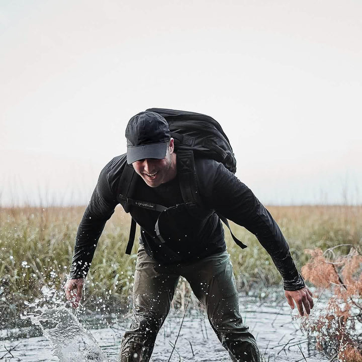 Man rucking through water in outdoor gear with GORUCK backpack, grassland background