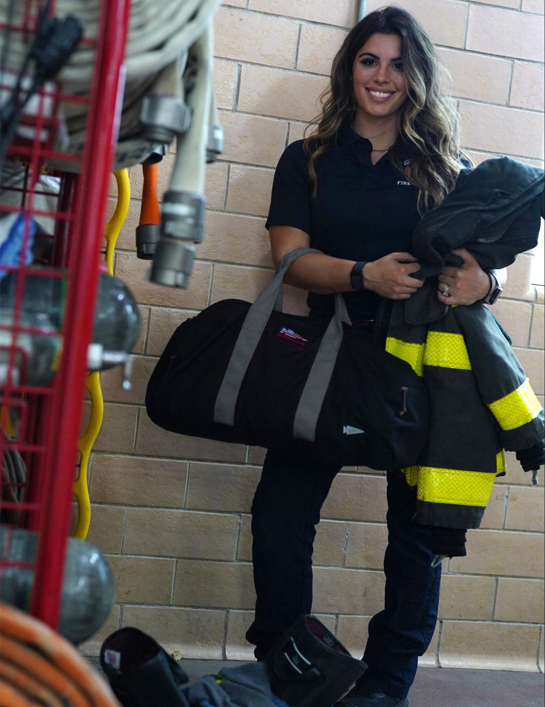 A smiling firefighter holding a black duffel bag and jacket stands next to firefighting equipment.