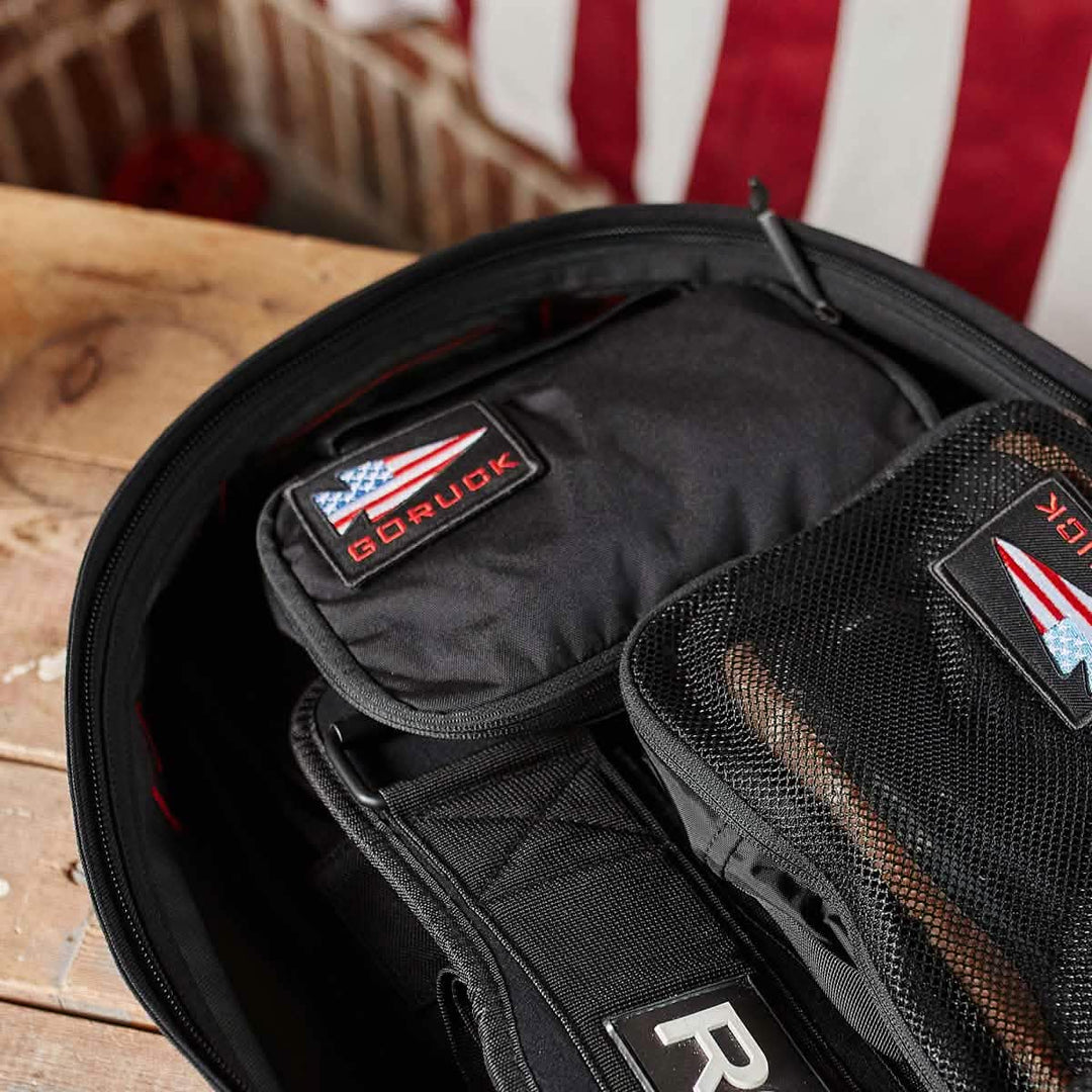 Open backpack with GORUCK Shadow Pockets made from durable Cordura, featuring American flag patch designs for organized storage, displayed on a wooden surface.