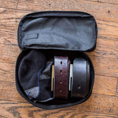 A GORUCK Shadow Pockets travel pouch sits open on a wooden floor, neatly holding two belts and a pair of socks inside.