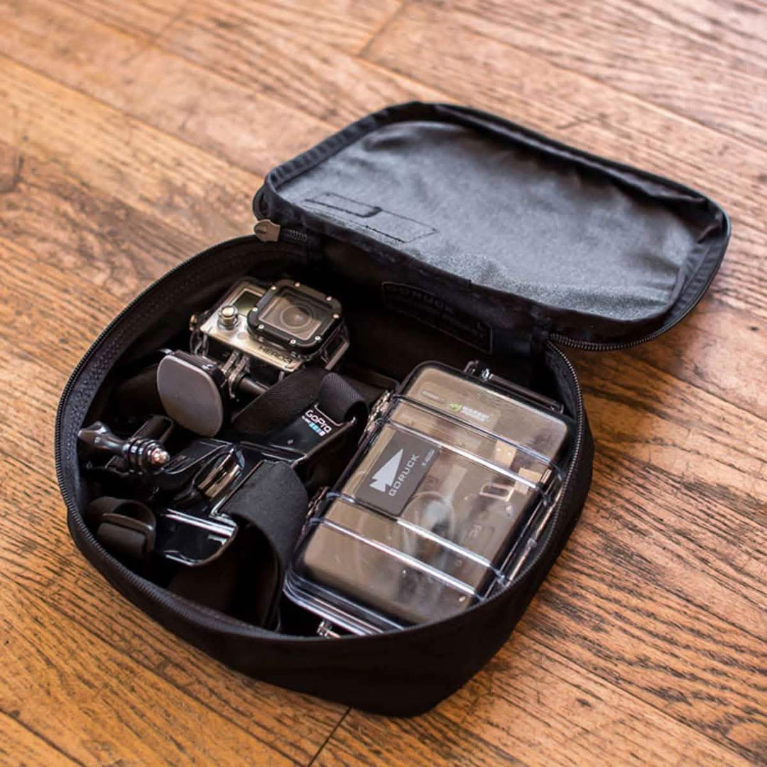 The GORUCK Shadow Pockets, a compact black Cordura case, holds your GoPro camera, gear, accessories, and a plastic storage box—everything organized on a wooden floor.