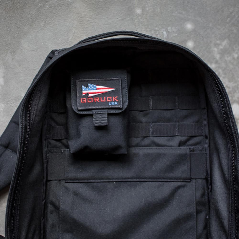 Open black GORUCK backpack with a visible USA patch inside on a textured surface, featuring MOLLE webbing for enhanced internal organization and equipped with the Simple Side Pocket.