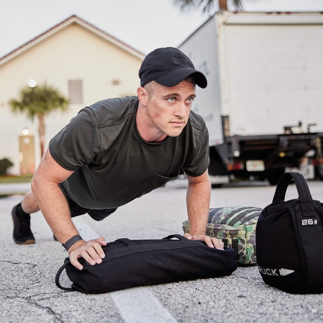 Man doing push-ups on pavement with GORUCK ruck gear, sandbags, and backpack outdoors