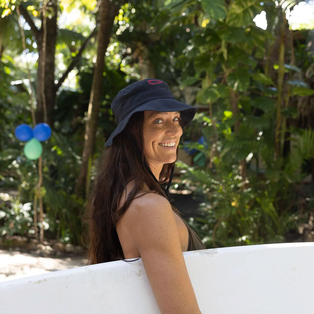 A woman with long dark hair in the GORUCK Boonie Hat - Slick - ToughDry holds a surfboard and smiles amid greenery and balloons. The hat’s ToughDry® fabric and 50+ UPF rating provide superior sun protection for outdoor adventures.