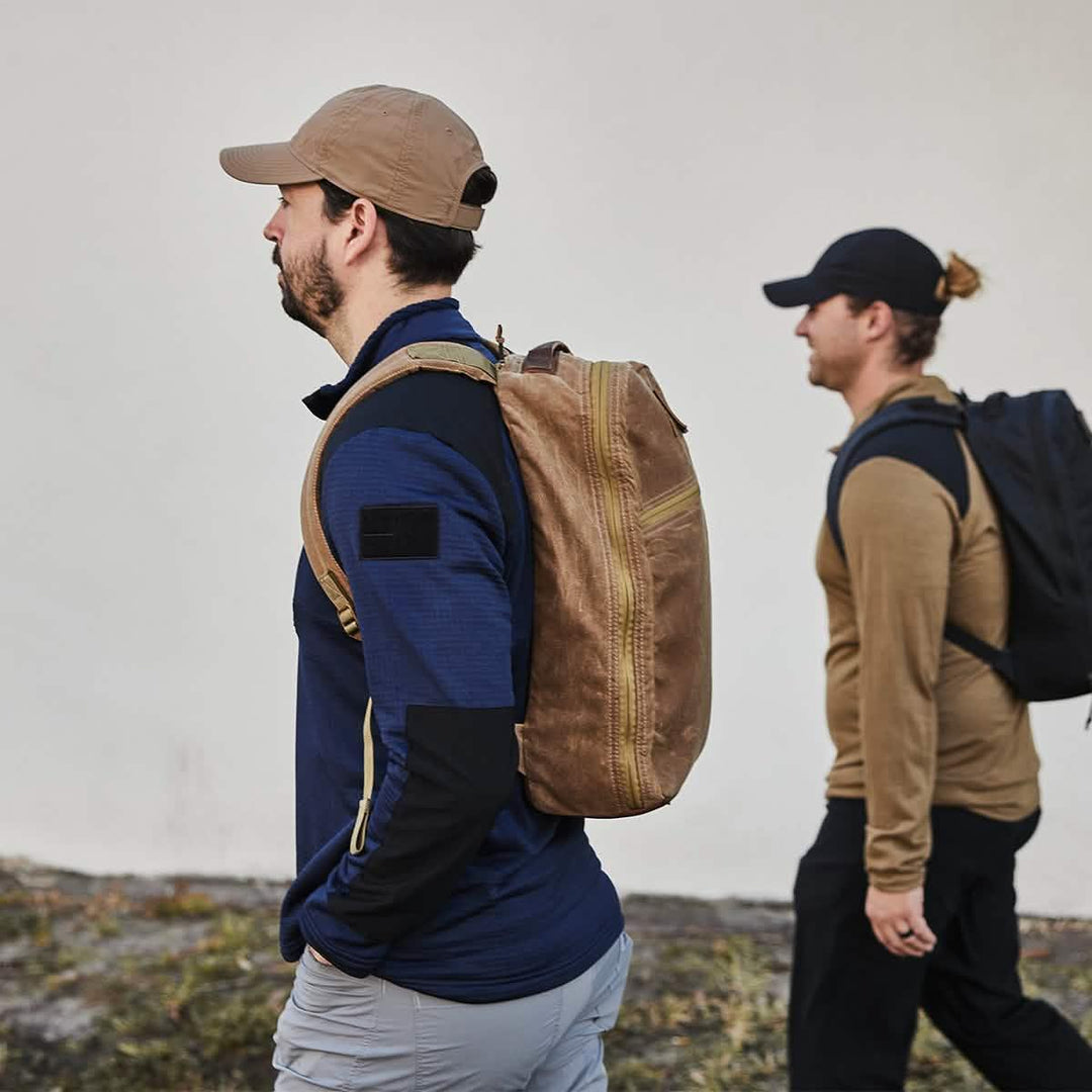 Two men rucking outdoors with GORUCK backpacks and hats against a neutral wall