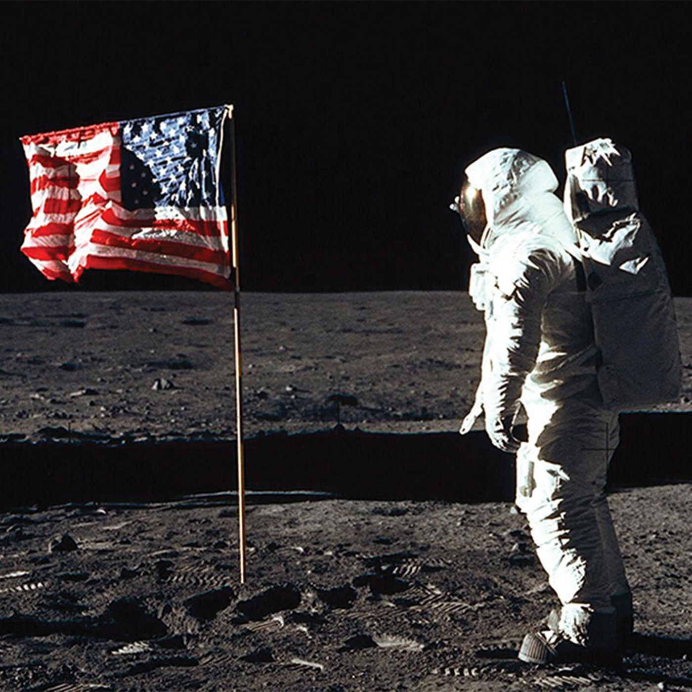 An astronaut in a spacesuit stands on the moon next to an American flag planted in the lunar soil.