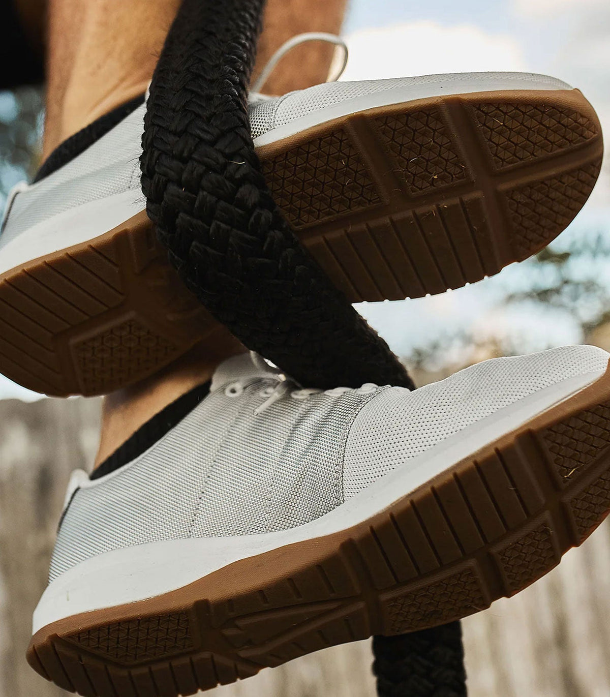 Close-up of a person wearing white sneakers with gum soles, climbing a thick black rope outdoors.
