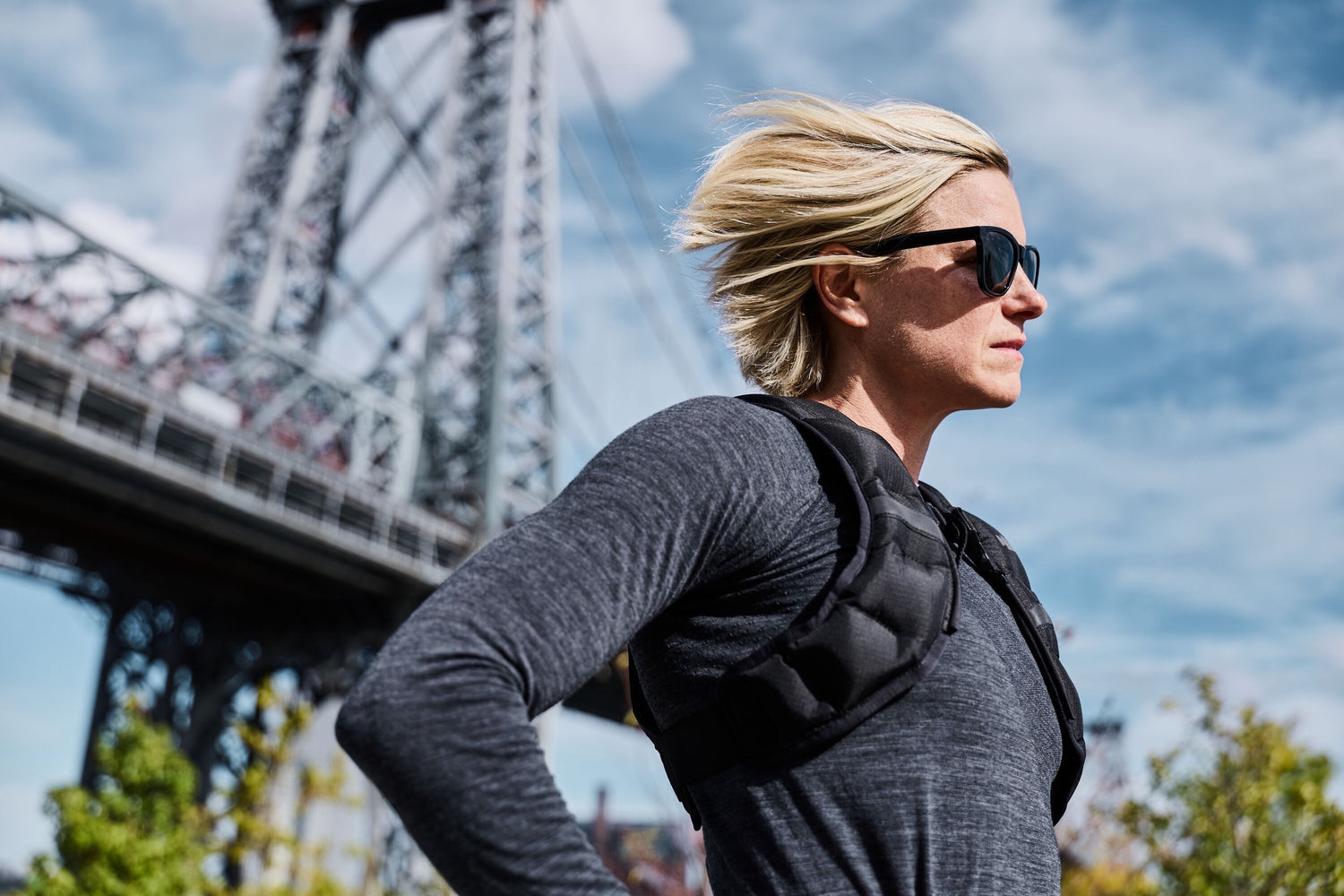 Person with short blond hair and sunglasses jogging near a large steel bridge on a sunny day.