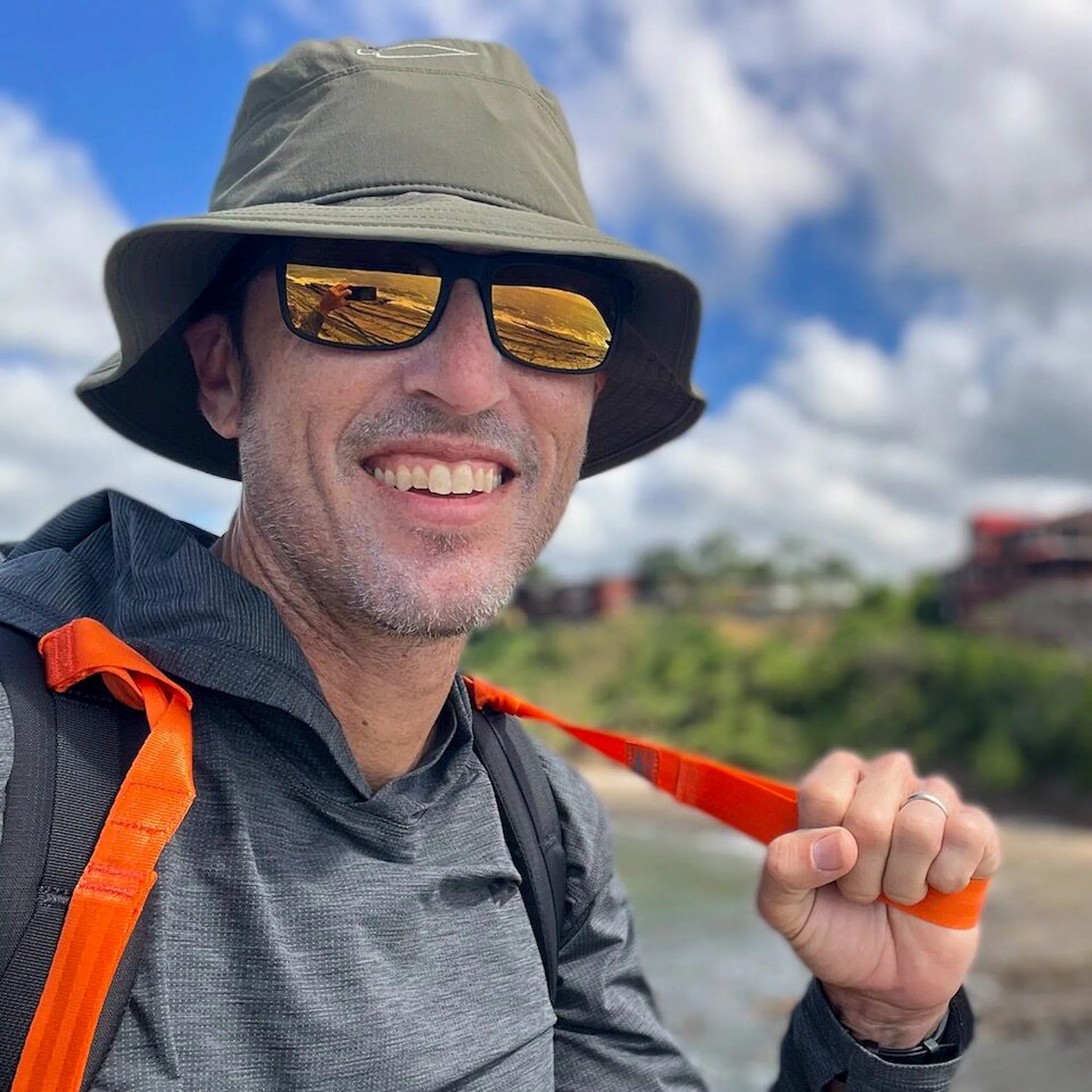 A smiling man in a hat and sunglasses holds a pair of SMC Gear T-REX Hand Straps outdoors, surrounded by greenery and sky—a great example of using rucking accessories to add comfort and help relieve hand swelling.