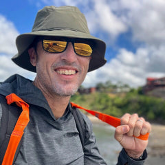 A smiling man in a hat and sunglasses holds a pair of SMC Gear T-REX Hand Straps outdoors, surrounded by greenery and sky—a great example of using rucking accessories to add comfort and help relieve hand swelling.