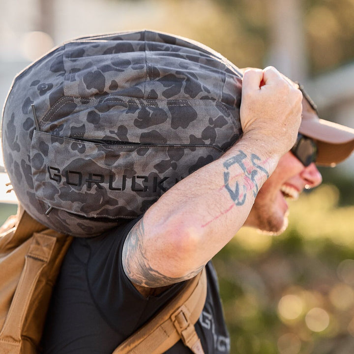 A smiling man carries a camo-patterned GORUCK Sand Medicine Ball on his shoulder outside, building core stability just as he would in his home gym.