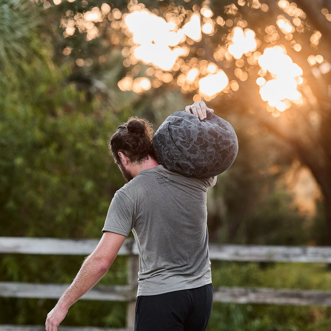 At sunset, a man carries a camo GORUCK bag—ideal for bringing his Sand Medicine Ball to train core stability after a home gym workout.