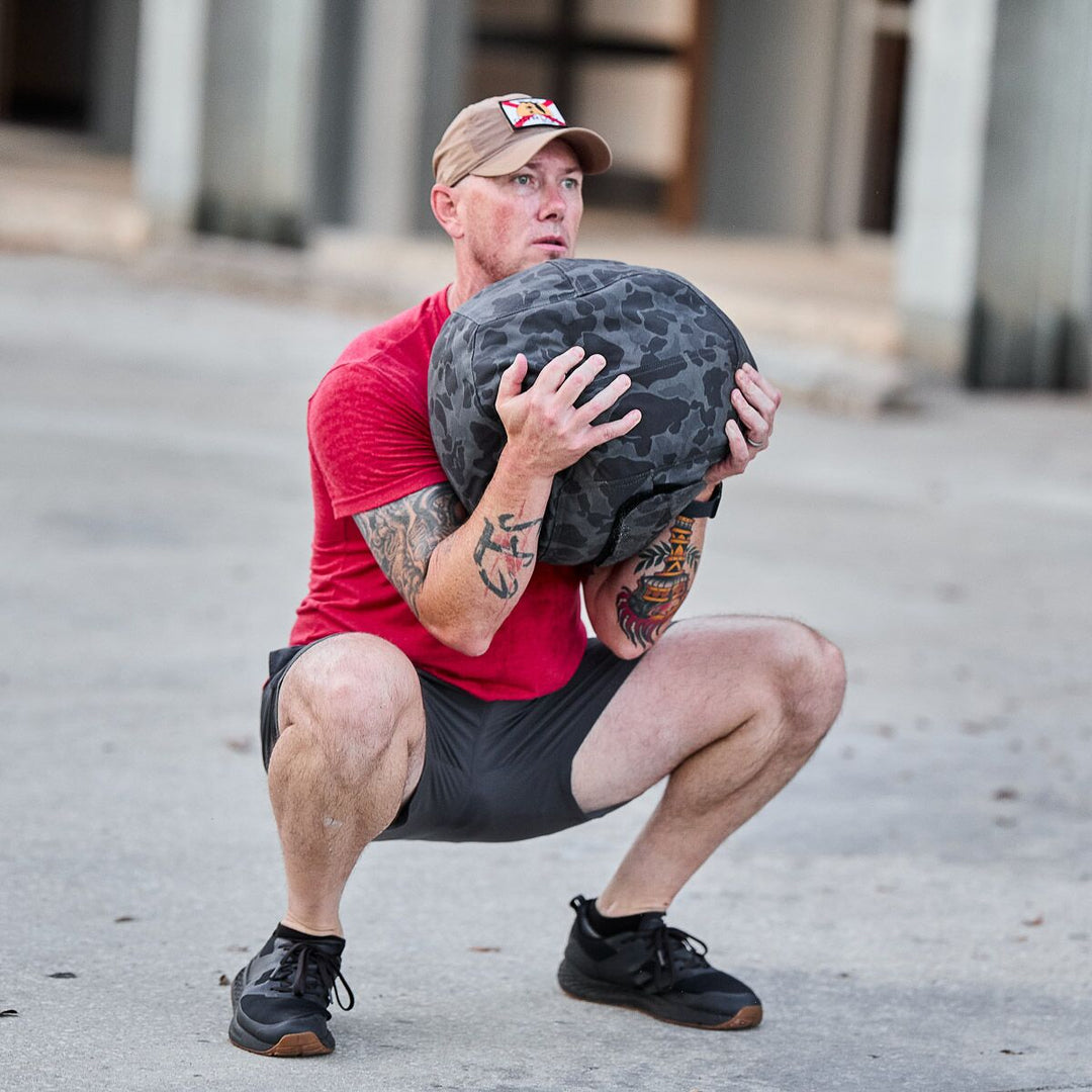 A man in a red shirt squats outdoors, holding the GORUCK Sand Medicine Ball at his chest to enhance core stability—ideal inspiration for your home gym workouts.