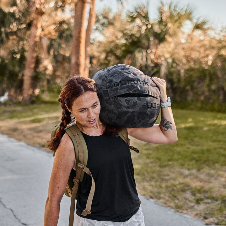 A woman with a braid carries a GORUCK Sand Medicine Ball on her shoulder outdoors, wearing a black tank top and backpack—showcasing the core stability often built through home gym workouts.