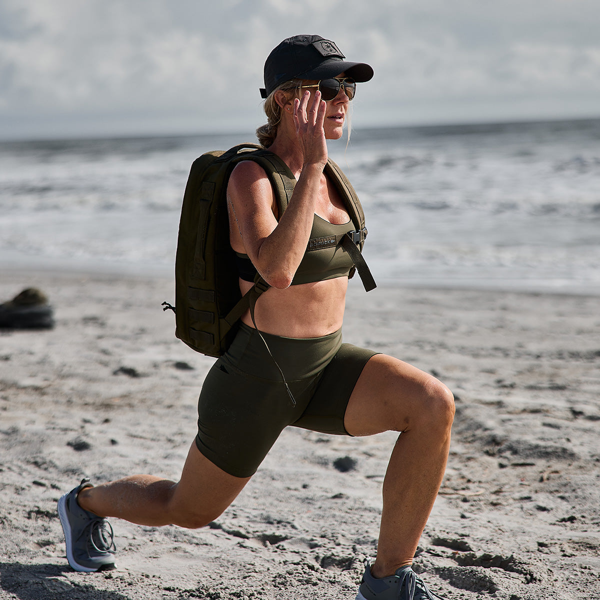 Woman in workout gear and cap lunging on a sandy beach, wearing a backpack, with ocean in the background.