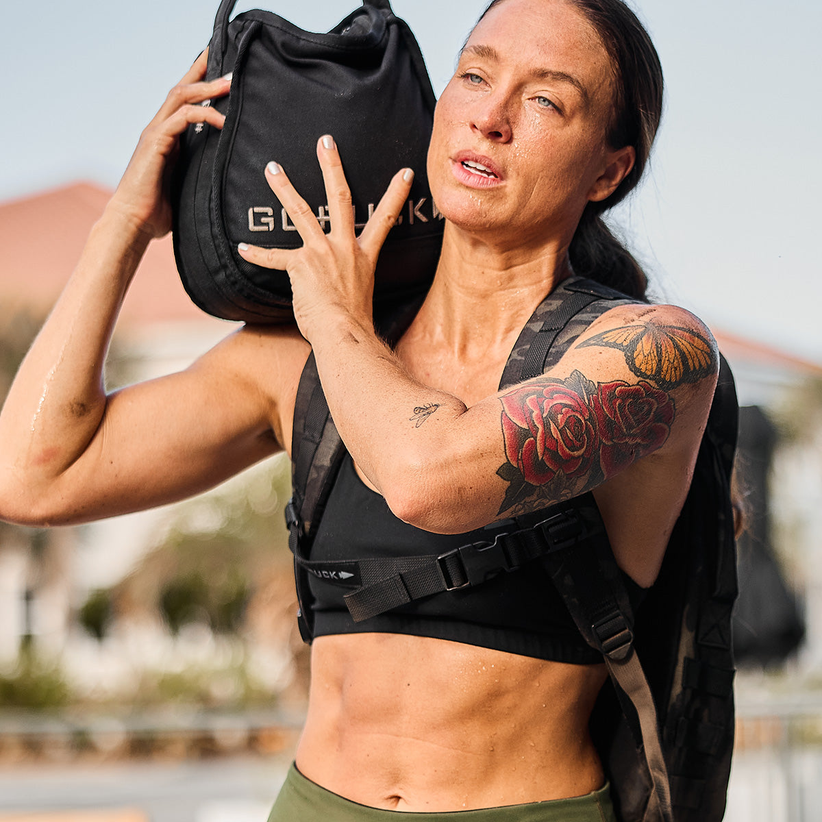Woman with a tattoo lifts a weighted bag outdoors, wearing a sports bra and backpack, looking focused.